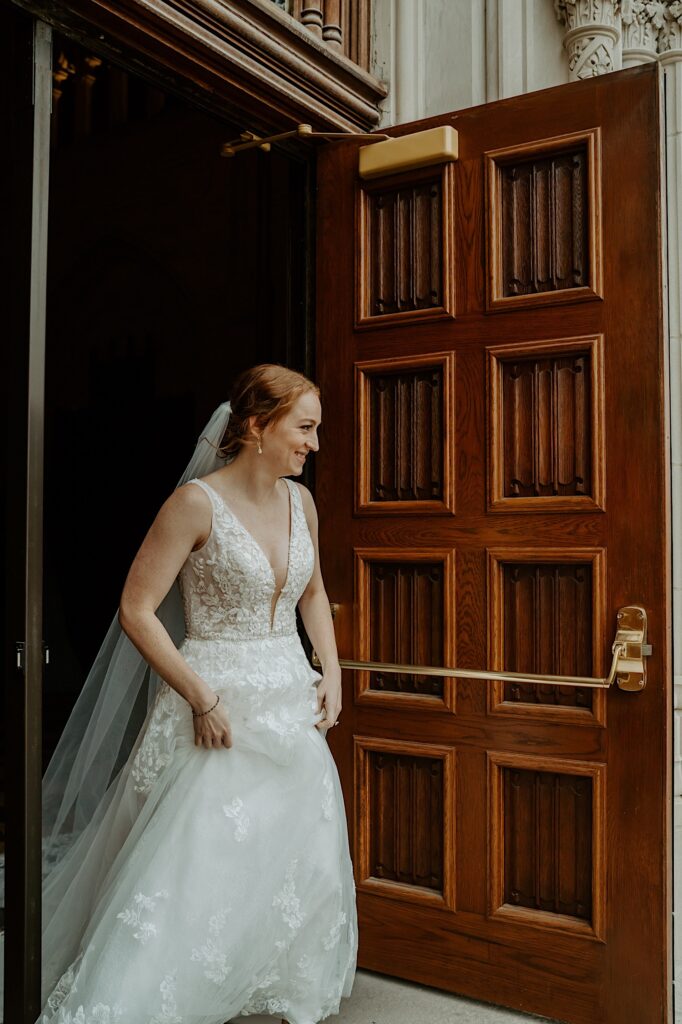 Bride in a lace deep v dress smiles as she walks out the church doors to greet her soon to be wife for a first look. 