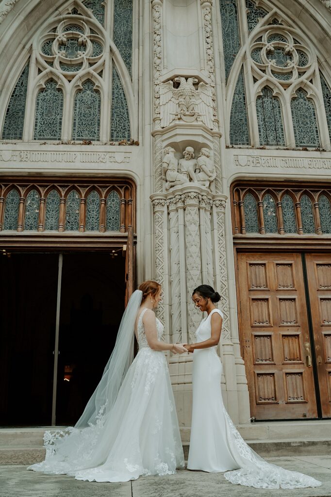 Brides stand holding hands in front of Scottish Rite Cathedral in Indianapolis before their wedding ceremony. 