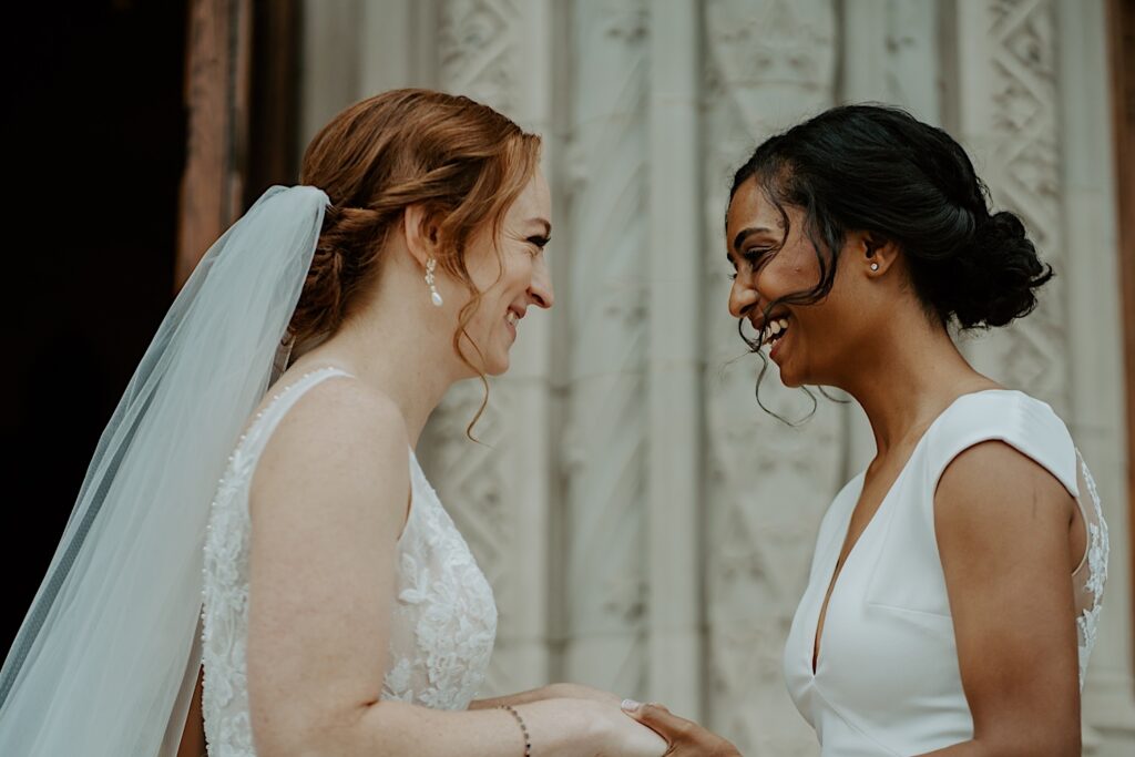 Brides hold each other's hands and laugh while looking into each other's eyes in front of the Scottish Rite Cathedral in Indianapolis. 