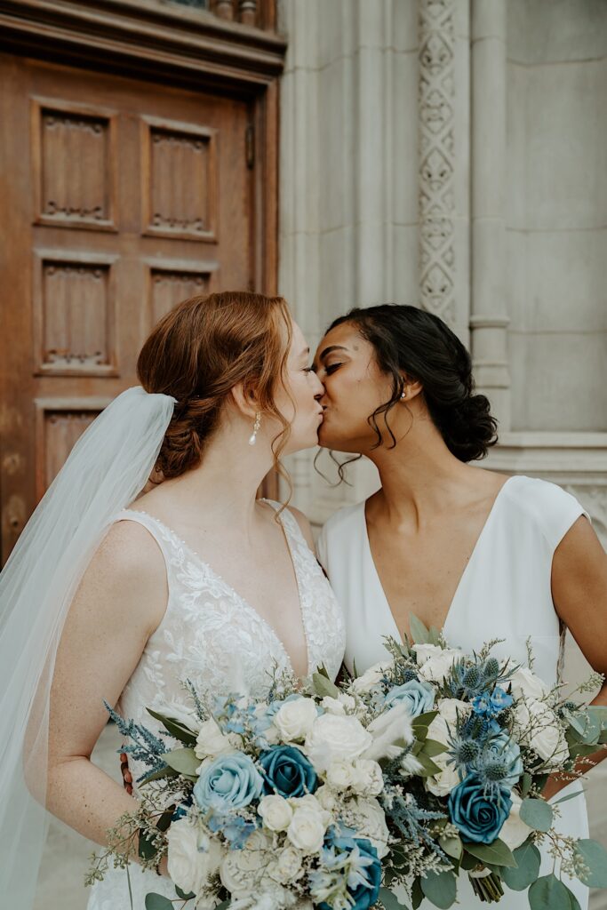 Brides lean in to kiss in front of Scottish Rite Cathedral in Indianapolis while holding gorgeous blue and white rose bouquets. 