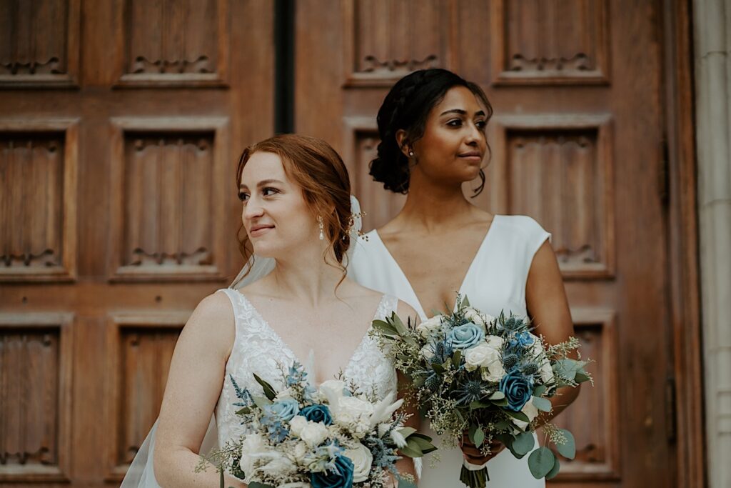 LGBTQ+ couple stands on the steps of Scottish Rite Cathedral holding white and blue bouquets and looking in opposite directions posing for Midwest Wedding Photographer. 