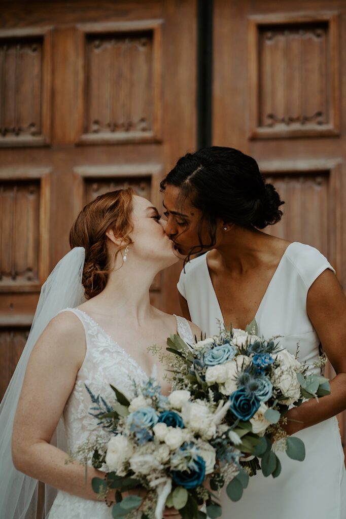 Brides lean in for a kiss on the steps of Scottish Rite Cathedral while holding blue and white rose bouquets. 