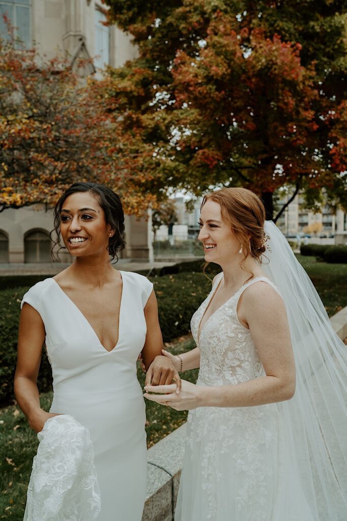 Brides stand under beautiful autumn trees and smile into the distance while holding hands. 