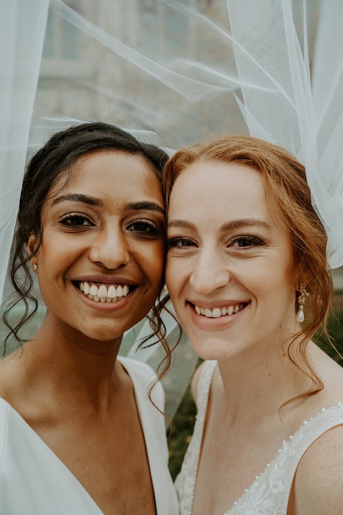 Brides smile and pose underneath one of their veils. 