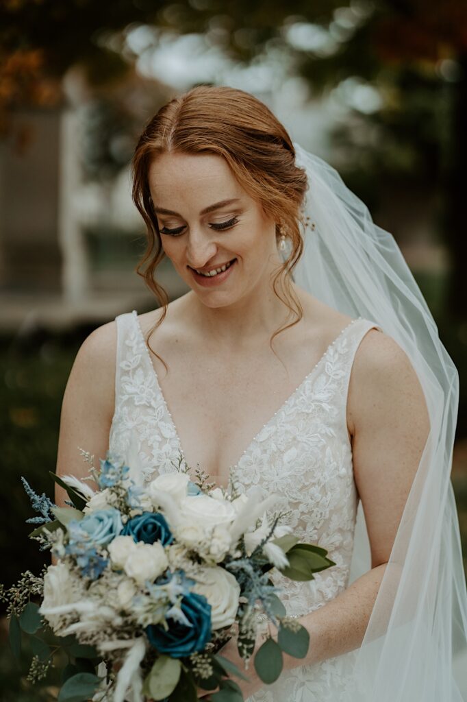 Bride in a lace dress with red hair looks down at her blue and white rose bouquet as her white veil is wrapped around her arm. 