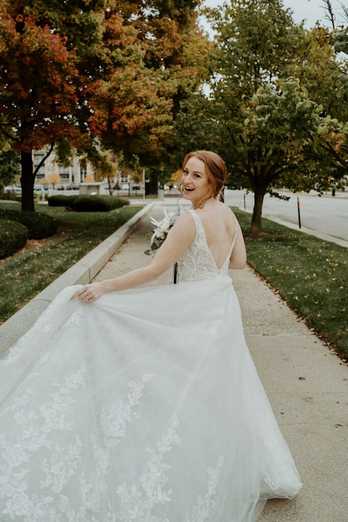 Bride in a lace dress runs down the sidewalk holding her skirt out and looking behind her at the camera.