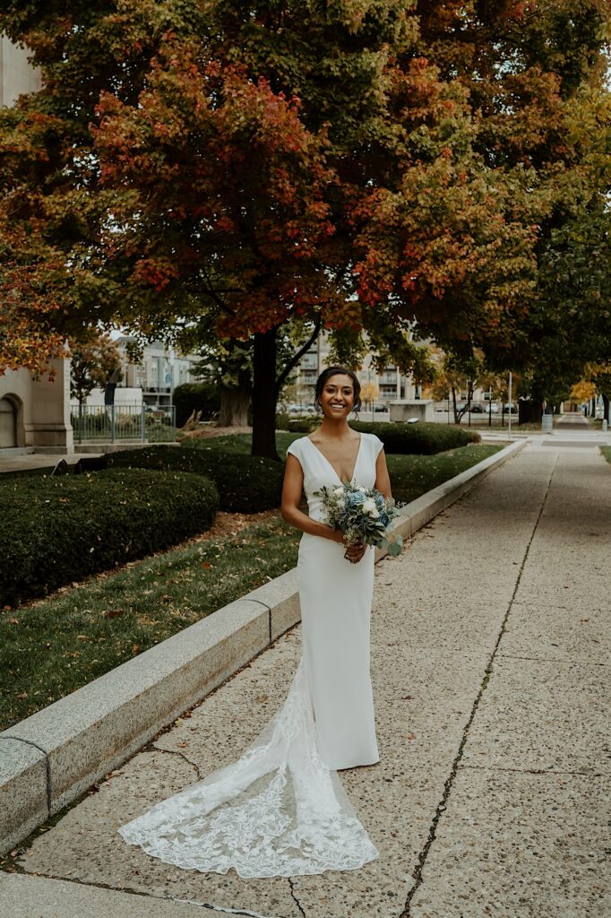 Bride stands on a sidewalk with autumn trees over her smiling at the camera for wedding portraits by Indianapolis Wedding Photographer. 