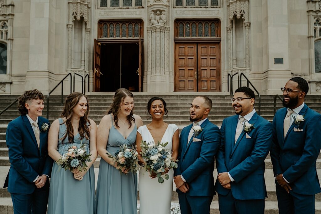 Bridal party stands around their bride laughing and smiling candidly in front of the cathedral in Indianapolis. 