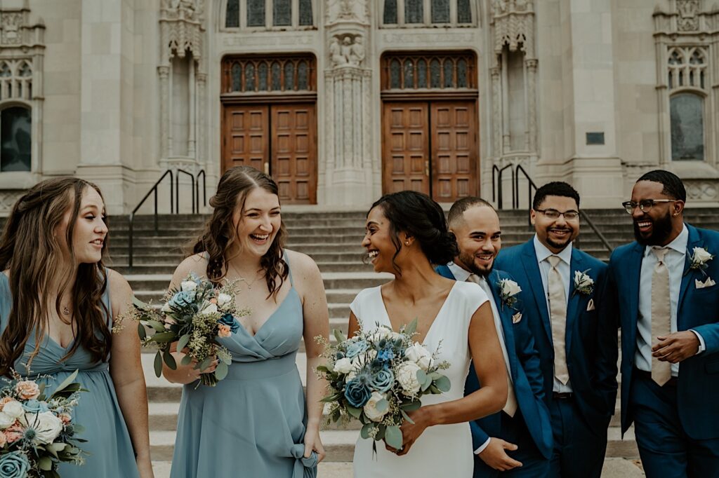 Bride holds her blue and white wedding bouquet and looks behind her shoulder at her bridesmaids as they walk towards the camera in front of the Scottish Rite Cathedral in Indianapolis. 