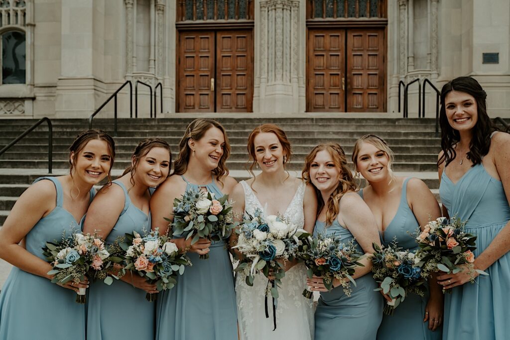 Bridesmaids holding colorful flower bouquets lean into bride and smile for the camera. 