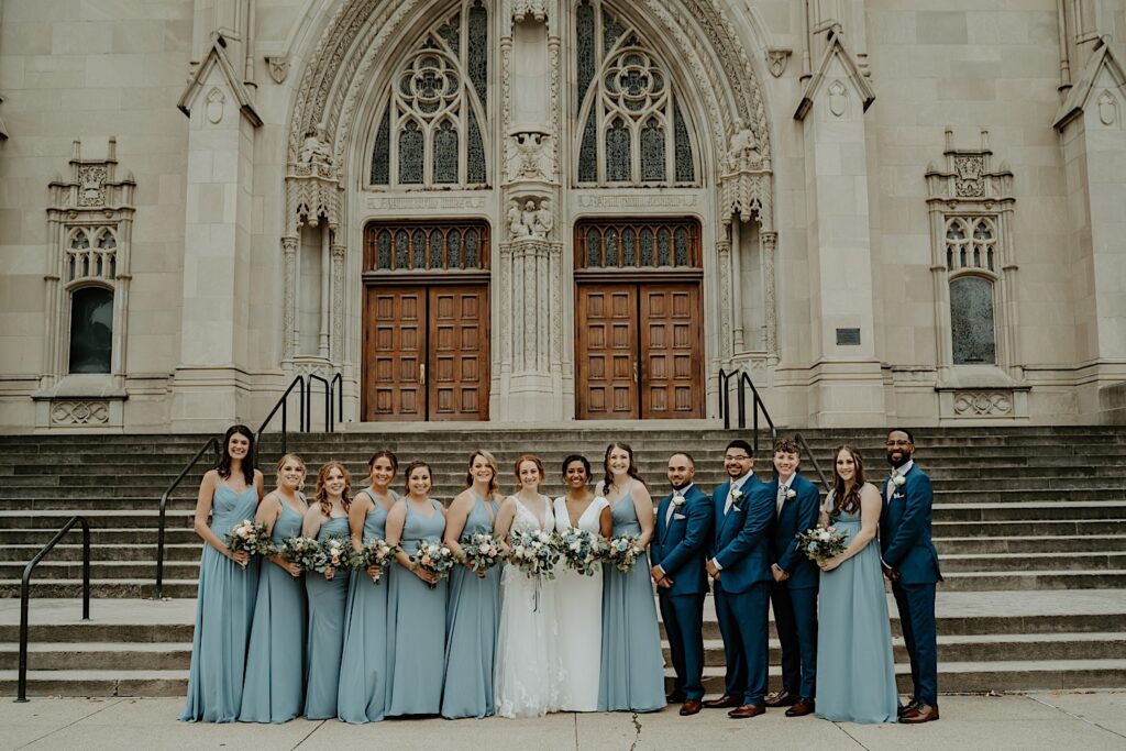 Wedding party in blue stands on both sides of the brides in white in front of the Scottish Rite Cathedral in Indianapolis. 