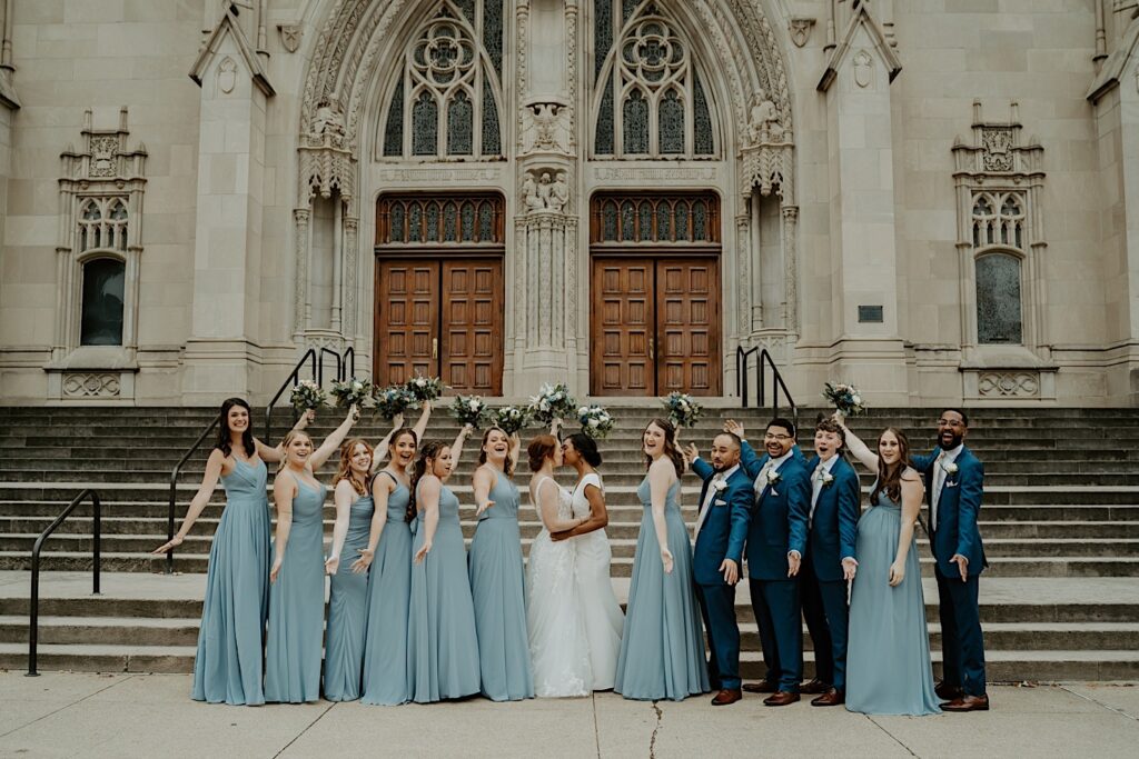 Wedding party in blue holds their bouquets and their arms up as their brides kiss and hold each other in the middle. They are all standing at the bottom step of the Scottish Rite Cathedral in Indianapolis. 