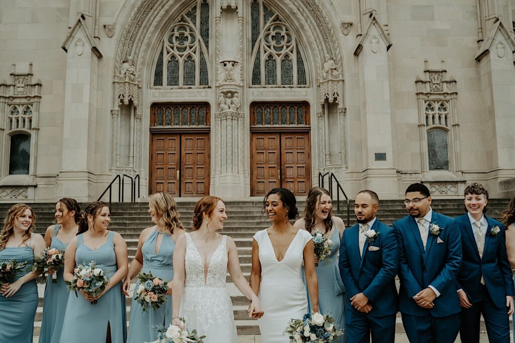 Brides look at each other while their wedding party stands around them smiling candidly for wedding portraits in front of the Scottish Rite Cathedral in Indianapolis. 