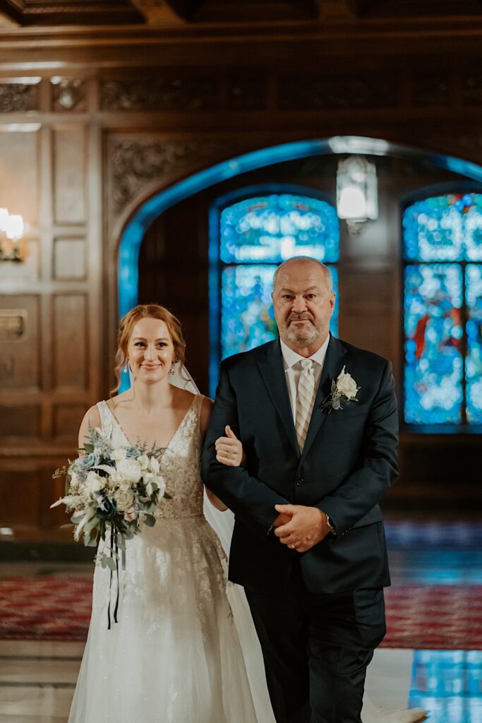 Bride in a lace dress holding a white and blue bouquet walks arm and arm with her father down the aisle at the Scottish Rite Cathedral in Indianapolis. 