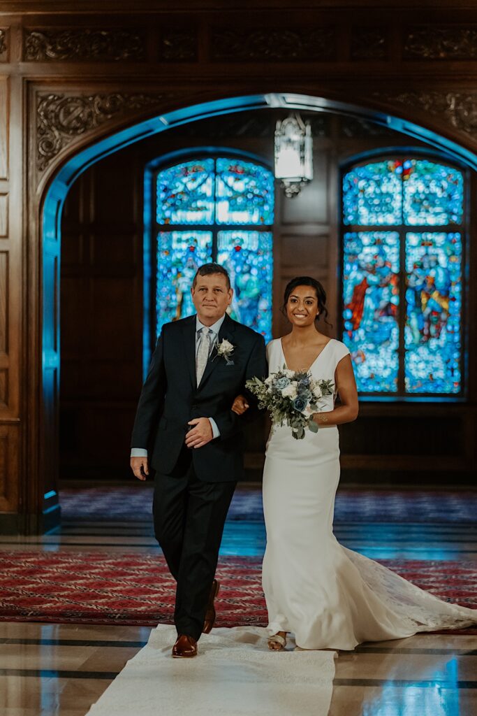 Bride in a minimalist wedding dress holding a white and blue bouquet walks arm and arm with her father down the aisle at the Scottish Rite Cathedral in Indianapolis. 