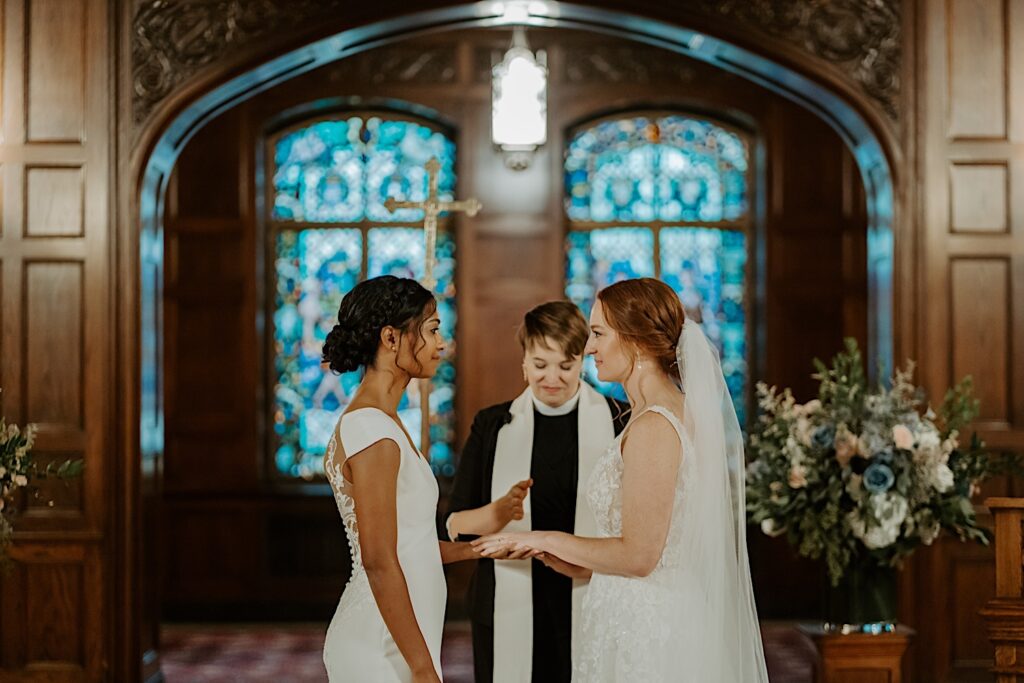 Brides look at each other at the end of the aisle in front of the stained glass at the Scottish Rite Cathedral in Indianapolis. 