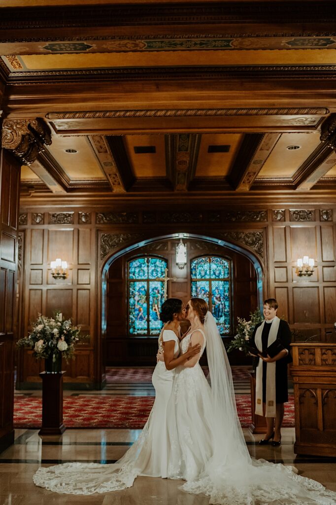 Brides kiss at the end of the aisle while their officiant stands on their right side and smiles in the Scottish Rite Cathedral in Indianpolis. 