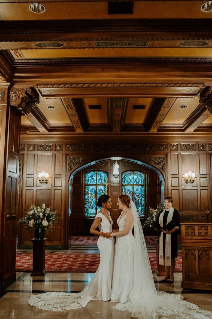 Brides look at each other after sharing a kiss with their officiant smiling while standing to their right in the Scottish Rite Cathedral in Indianapolis. 