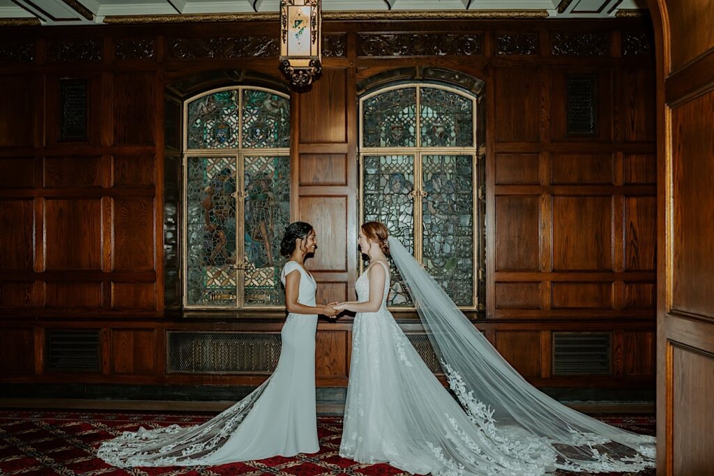  Brides hold hands and look at each other for a posed wedding portrait in front of stained glassed windows at oak walls in the Scottish Rite Cathedral.