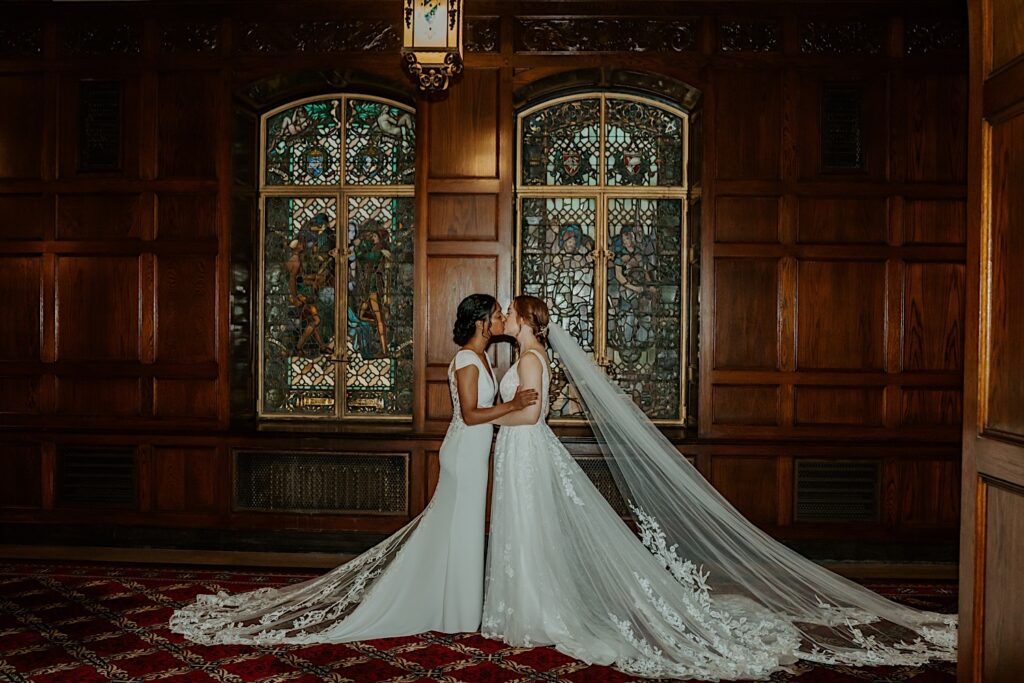 Brides kiss for a posed wedding portrait in front of stained glassed windows at oak walls in the Scottish Rite Cathedral.