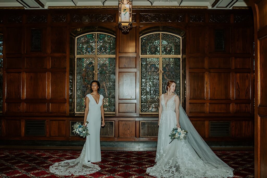 Brides stand 5 ft apart and look over their shoulders for a posed wedding portrait in front of stained glassed windows at oak walls in the Scottish Rite Cathedral. 