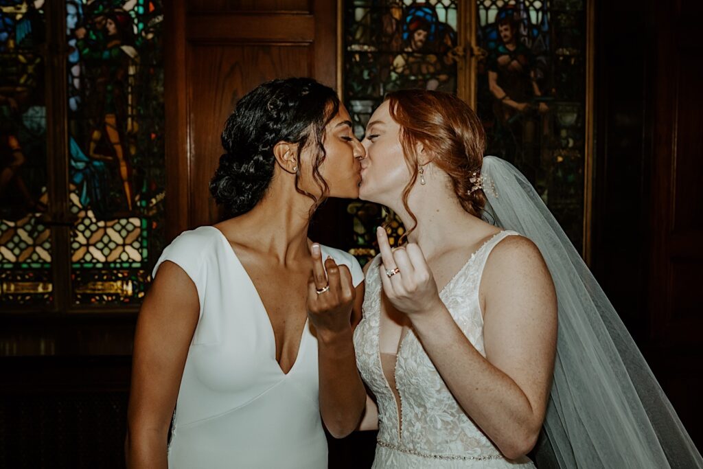 Brides kiss while holding their ring fingers to the camera against stained glass windows in the background at Scottish Rite Cathedral. 