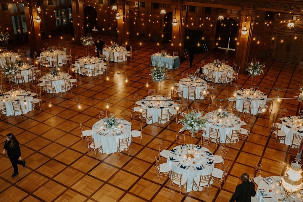 Indianapolis Wedding Photographer captures reception room from the balcony showcasing circle reception dinner tables and string lights on a wooden checkerboard floor. 