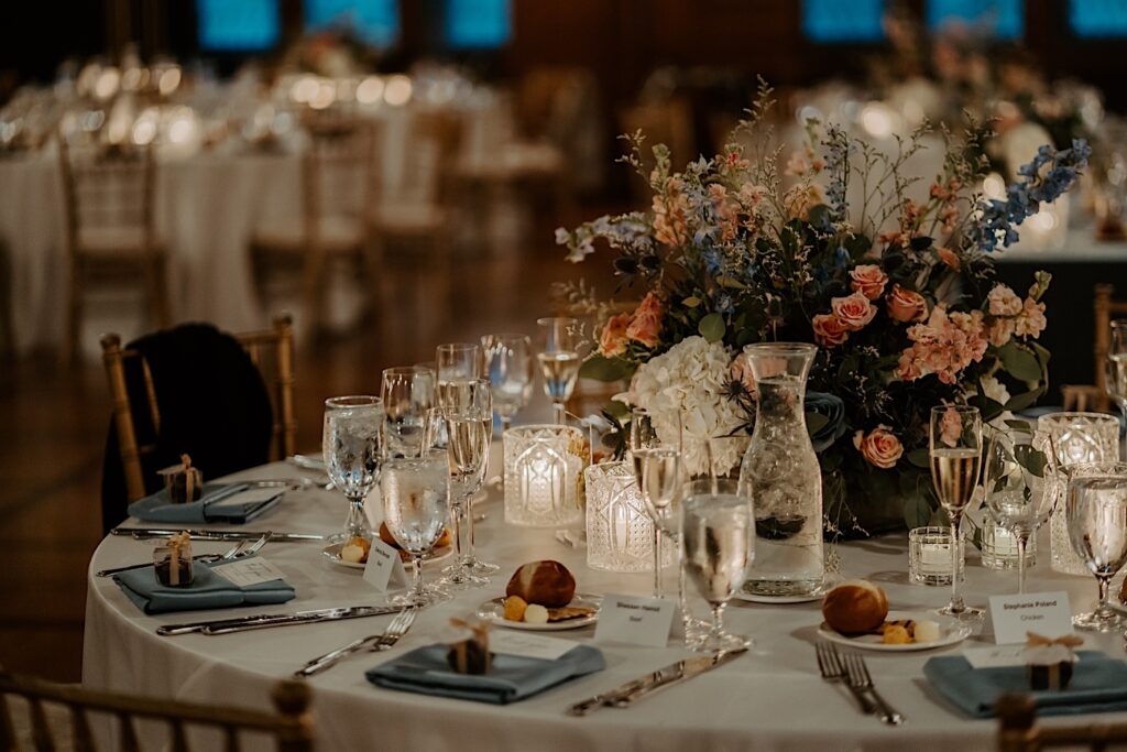 White table holds 8 table settings with silverware, teal napkins, bread plates, ice water glasses, champagne glasses, candles and large floral bouquet in the center. 