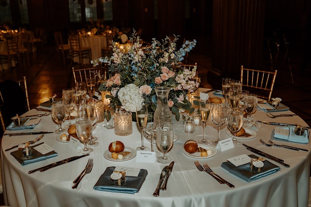 White table holds 8 table settings with silverware, teal napkins, bread plates, ice water glasses, champagne glasses, candles and large floral bouquet in the center. 