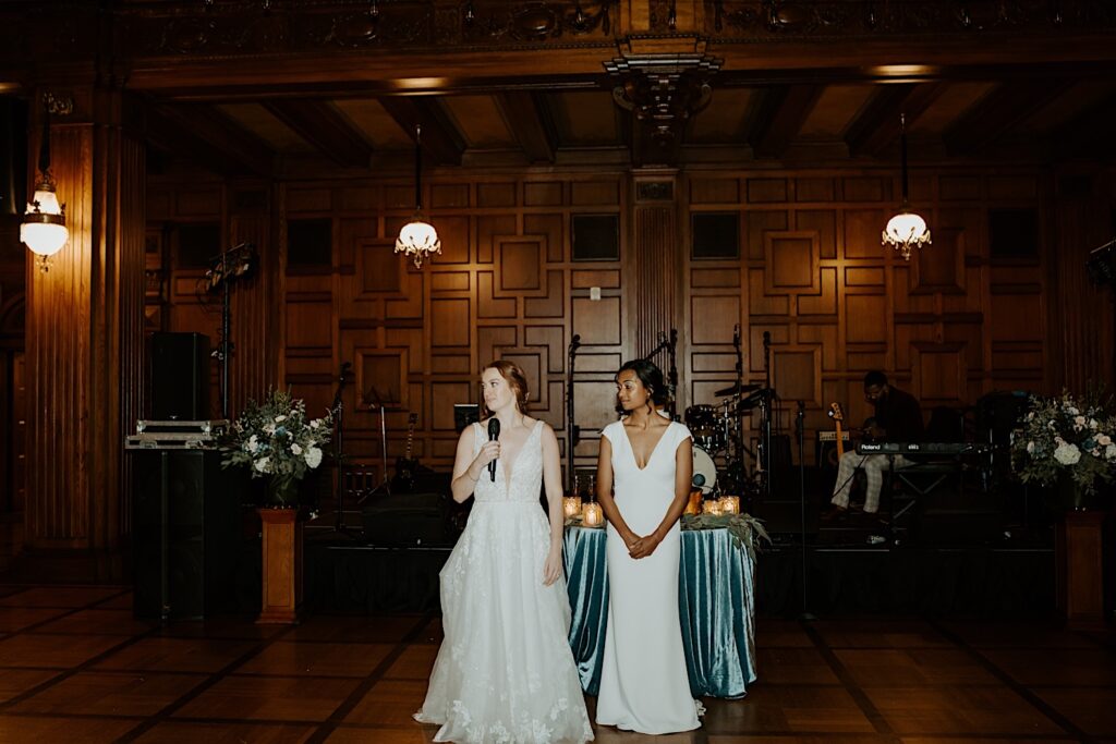 Brides stand in front of their head table as the bride on the left holds a microphone and welcomes their guests to their wedding reception. 