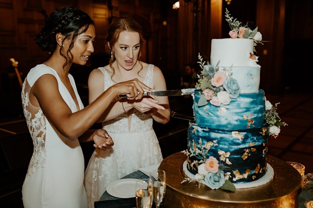 Brides share the cake knife as they cut a 4 teared blue and white cake with beautiful flowers stuck in every tier. 