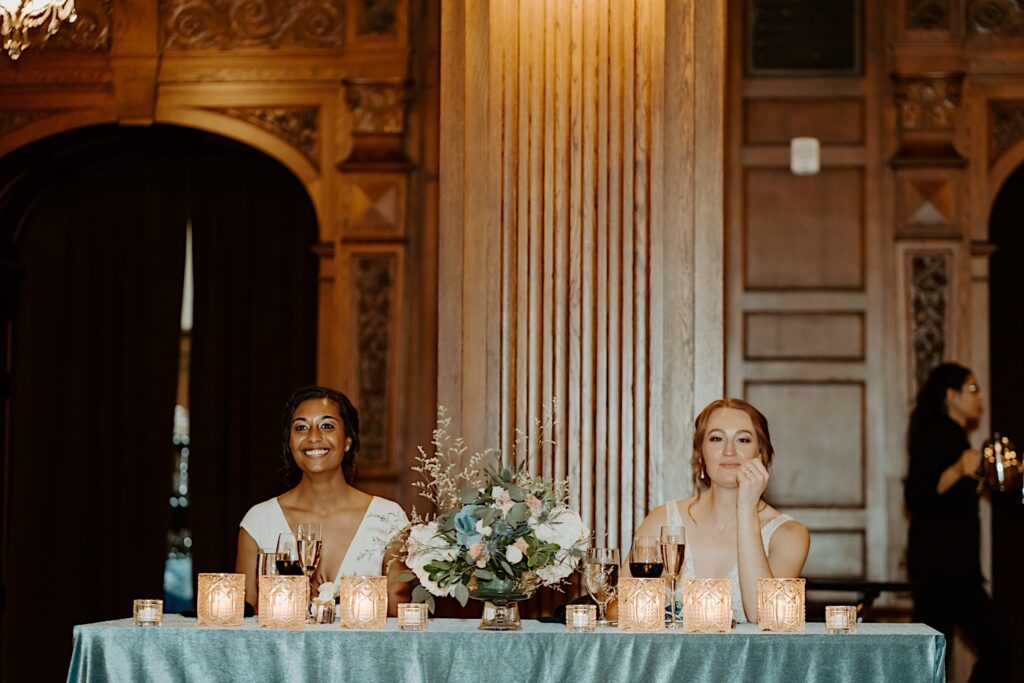 Bride's sit at the head table adorned with candles, and a flower bouquet with a teal blue tablecloth and smile at their guests during the reception at Scottish Rite Cathedral in Indianapolis.