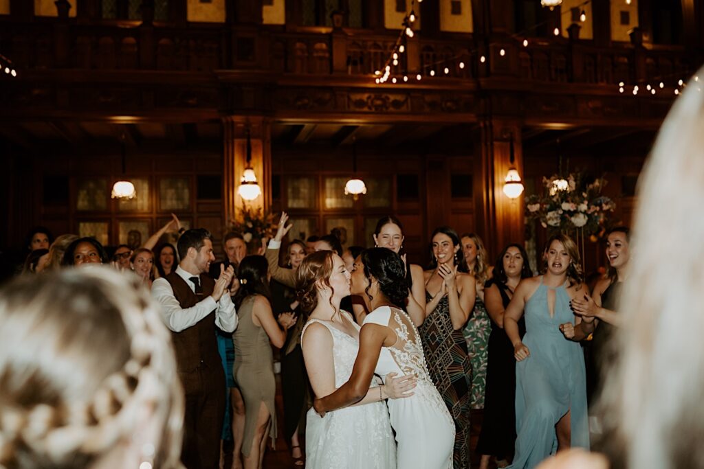 Brides kiss in the middle of the dance floor during wedding reception as guests clap and cheer around them. 