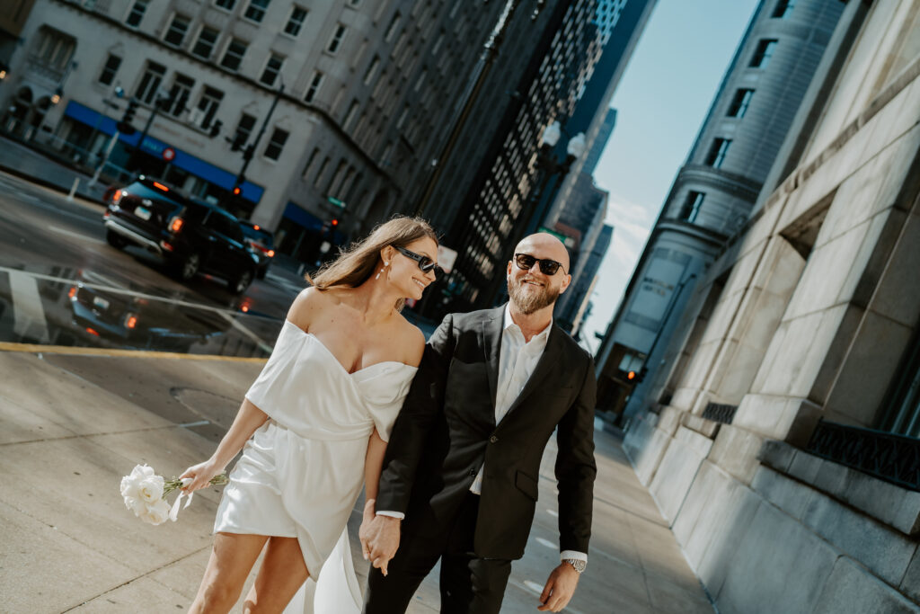 A woman in a slouchy of the shoulder white dress and black sunglasses walks while looking at her fiancé in a black suit down the streets of Chicago smiling. 