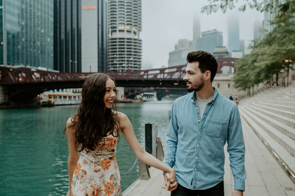 Woman in an orange floral dress holds hands with her fiancé in a denim button up as they walk along the Chicago Riverwalk with the river and bridges and downtown Chicago buildings in the background. 