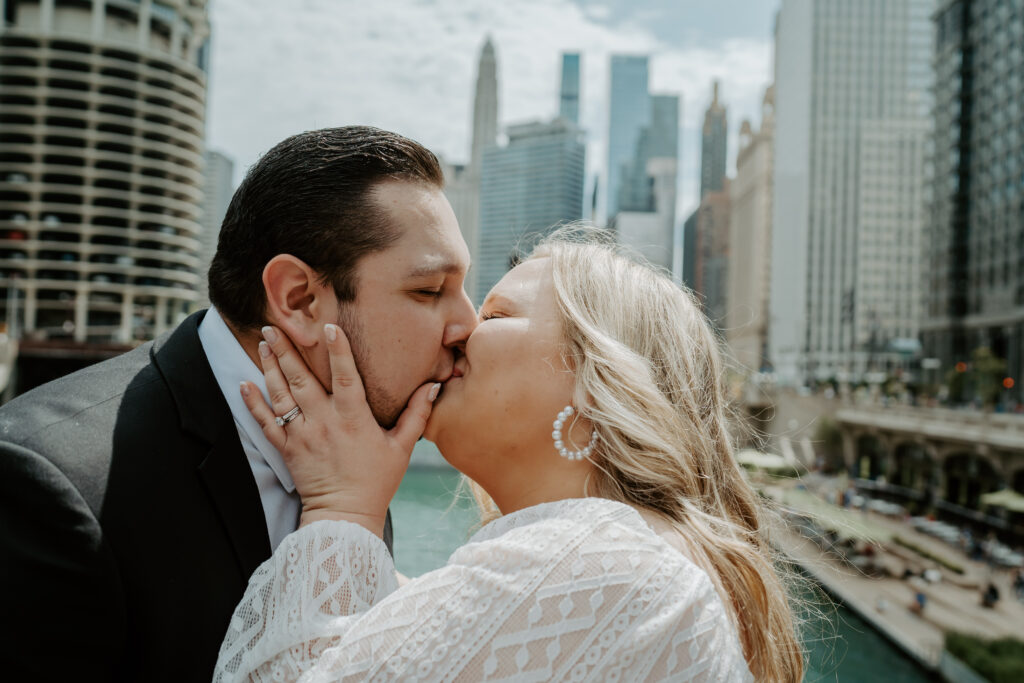 Couple kisses on a bridge overlooking the Chicago river with the city behind them for gorgeous Chicago engagement portrait.s 