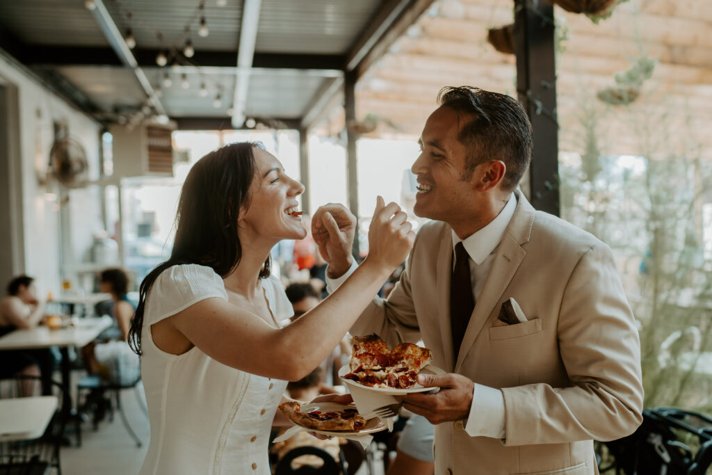 Woman in a white dress holds a plate of pizza and feeds her fiancé a bite with a fork as he does the same to her. They stand on the patio of a restaurant with patrons eating in the background. 