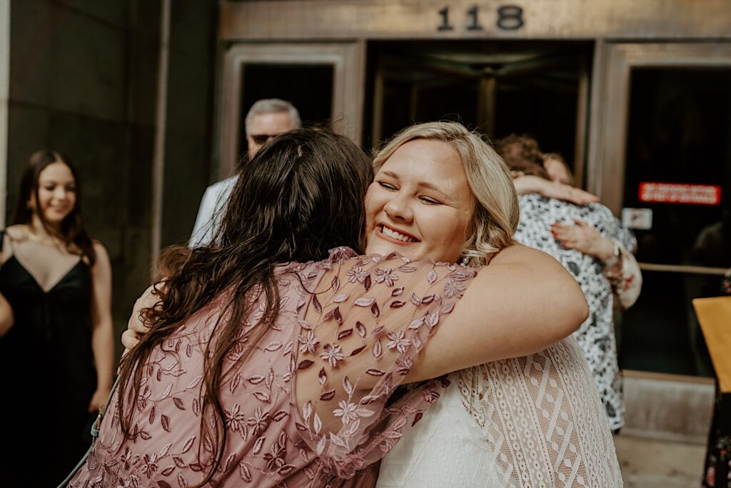 Bride in a white lace dress hugs her sister in a mauve dress in front of Chicago City Hall before their elopement ceremony. 