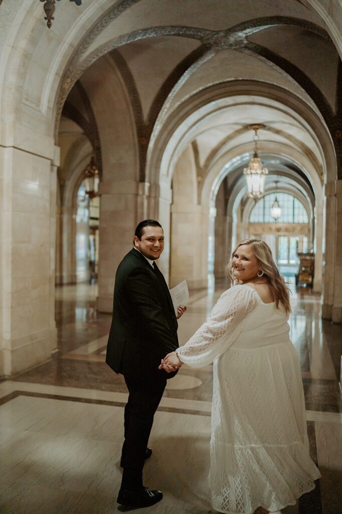 Bride and groom turn to face the camera as they walk down City Hall Hallway before elopement ceremony in Chicago. 