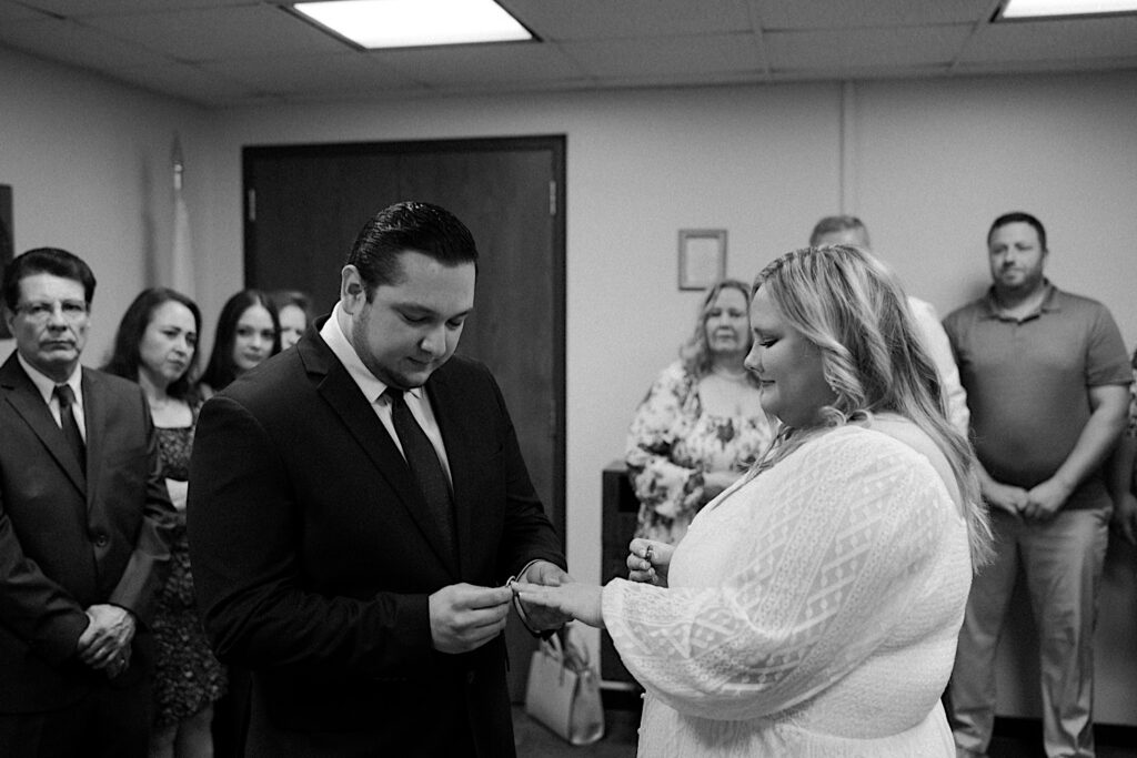 Groom puts ring on bride's finger during elopement ceremony ring exchange at Chicago City Hall as their families stand behind them.  