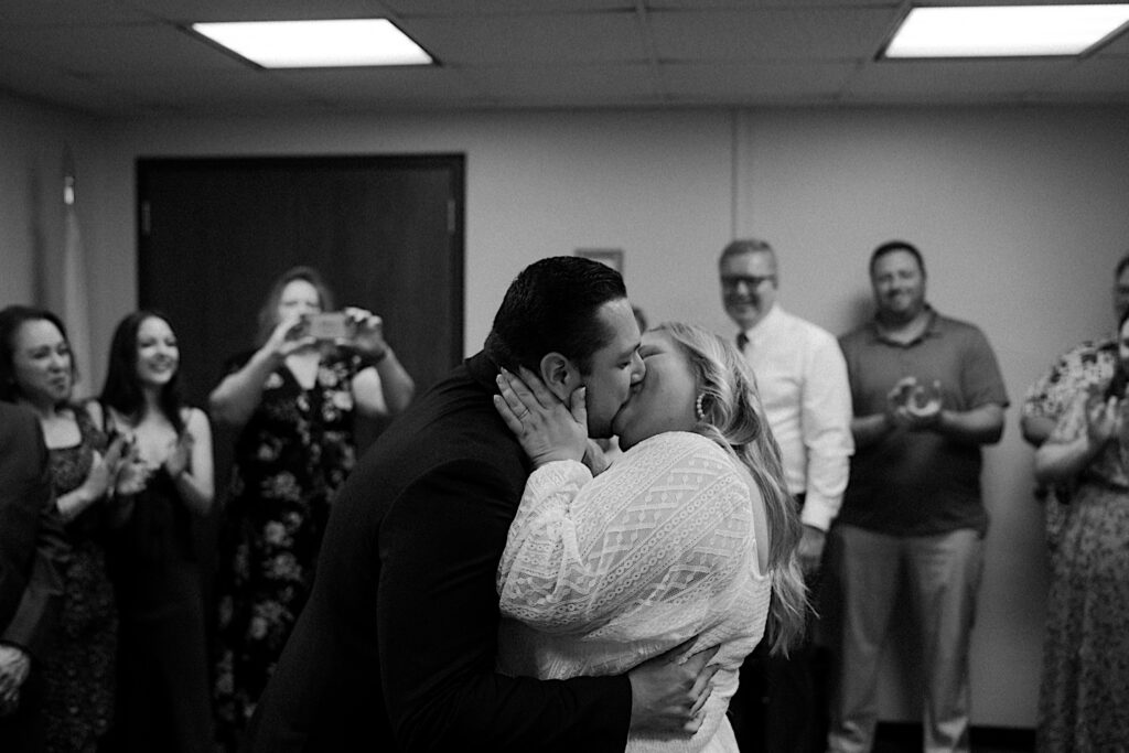 Bride holds groom's neck as they kiss during intimate elopement ceremony while their families clap behind them. 