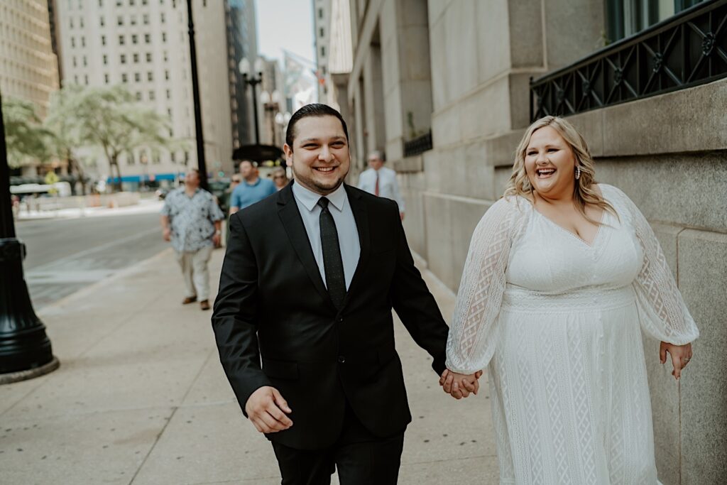 Bride in a long sleeve relaxed dress and Groom walk hand and hand down Chicago streets outside City Hall after wedding elopement ceremony. 