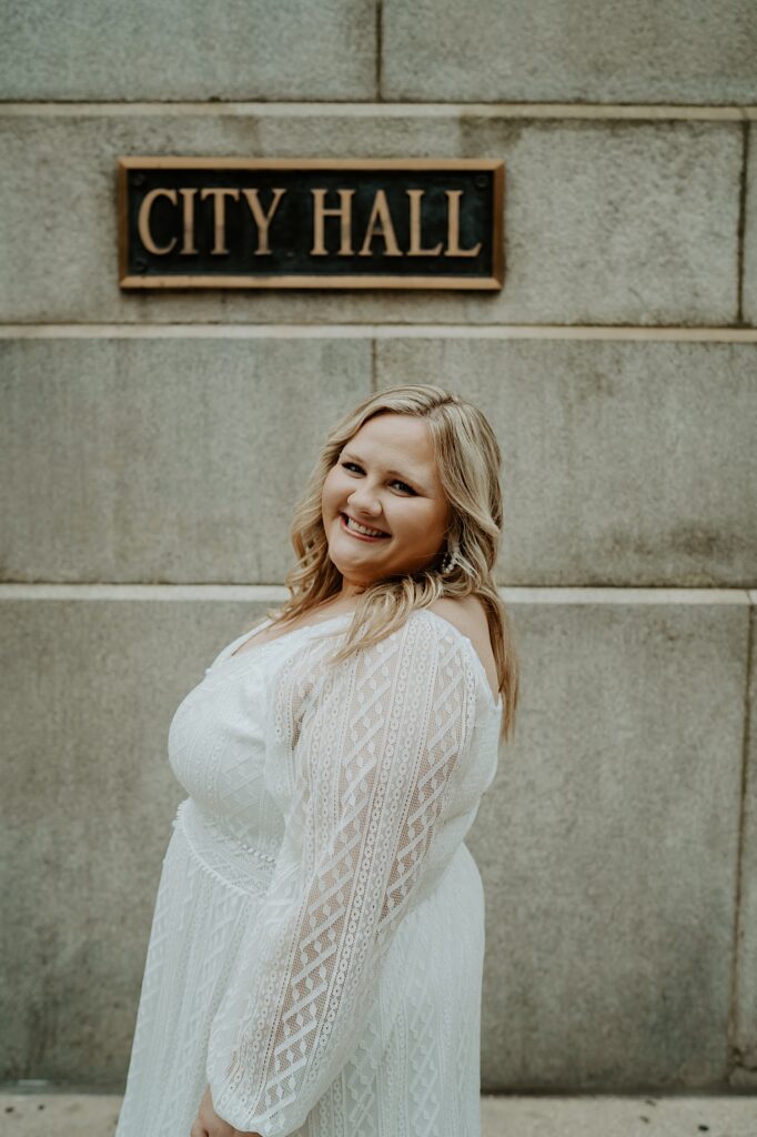 Bride in a relaxed white dress smiles at the camera next to City Hall sign at Chicago's City Hall after elopement ceremony. 