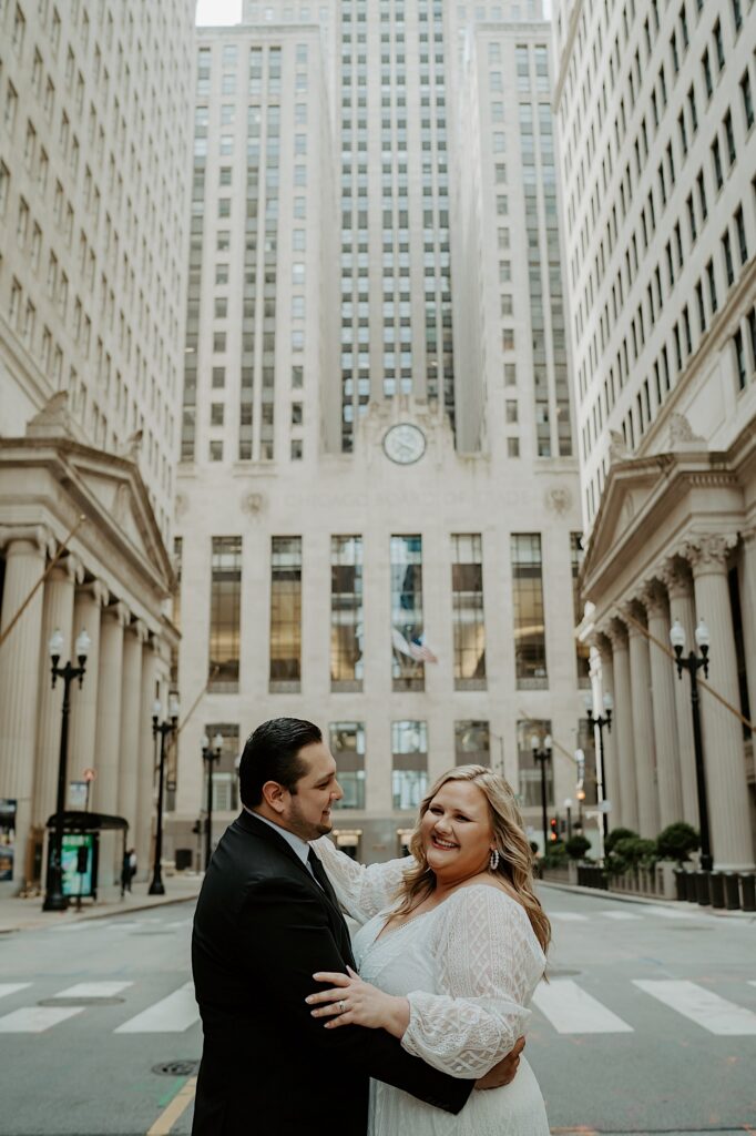 Bride looks towards the camera as the Groom looks towards her as they stand centered in the street with the Chicago Board of Trade in the background. 