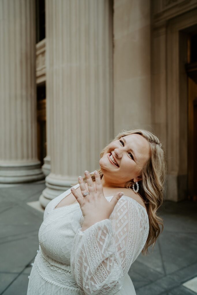 Bride flips her hair back and shows off her left hand with her diamond engagement ring while standing in front of a large columned building in downtown Chicago. 