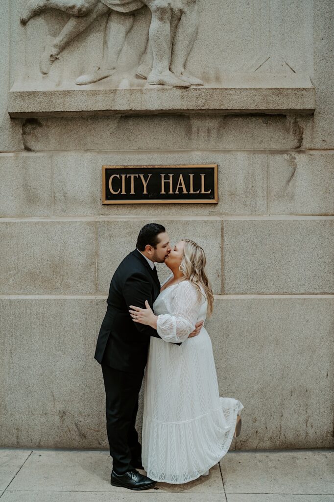 Bride lifts up her foot as Groom kisses her in front of City Hall sign. 