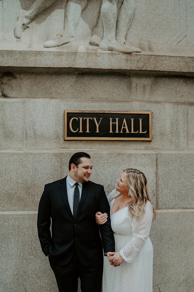 Bride holds onto Groom's arm as they look at each other underneath the City Hall sign for elopement portraits in Chicago. 