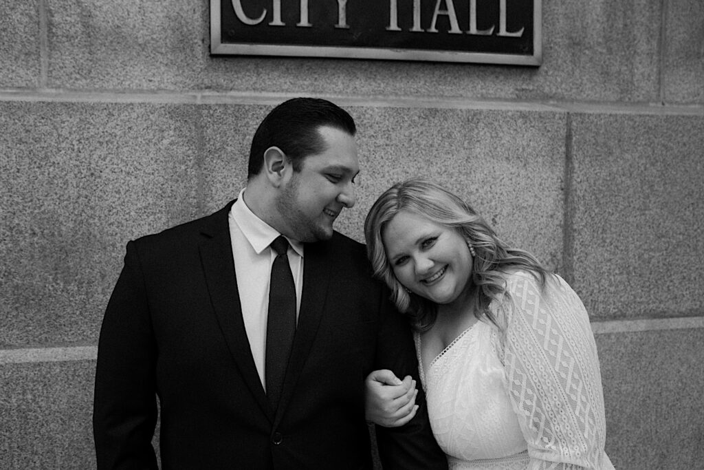 Bride looks into the camera as the Groom looks at her and they pose in front of a brick wall in a stunning black and white portrait. 