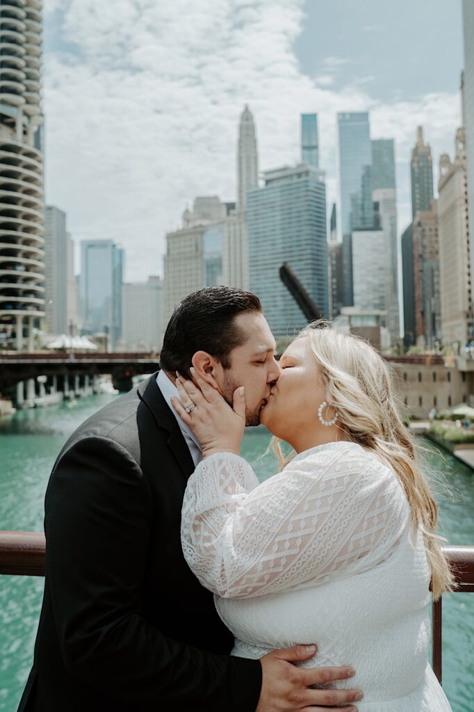 Bride and Groom embrace and kiss as they stand on a bridge overlooking the Chicago River with Chicago skyline in the background. 