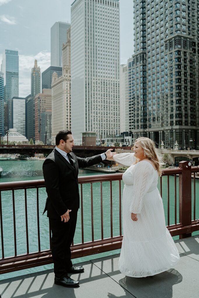 Groom holds his wife's arm as they stand on Chicago bridge overlooking the Chicago river with Wacker Drive buildings in the background. 